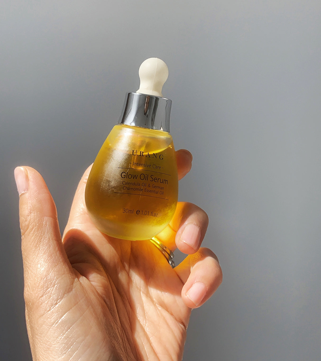 Hand holding the Urang Glow Oil Serum, showing the bright golden-yellow liquid being dispensed from a dropper.