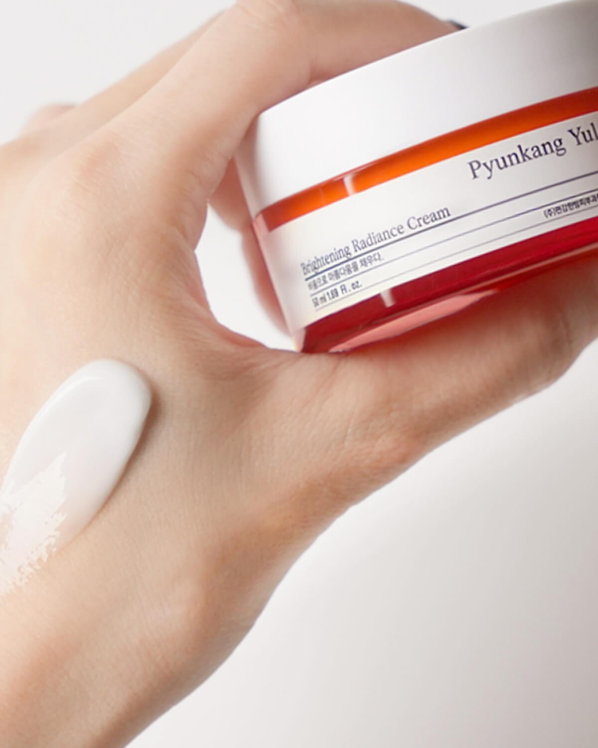 A close-up of a hand holding the Pyunkang Yul Brightening Radiance Cream jar, with a generous swipe of the white cream visible on the back of the hand.