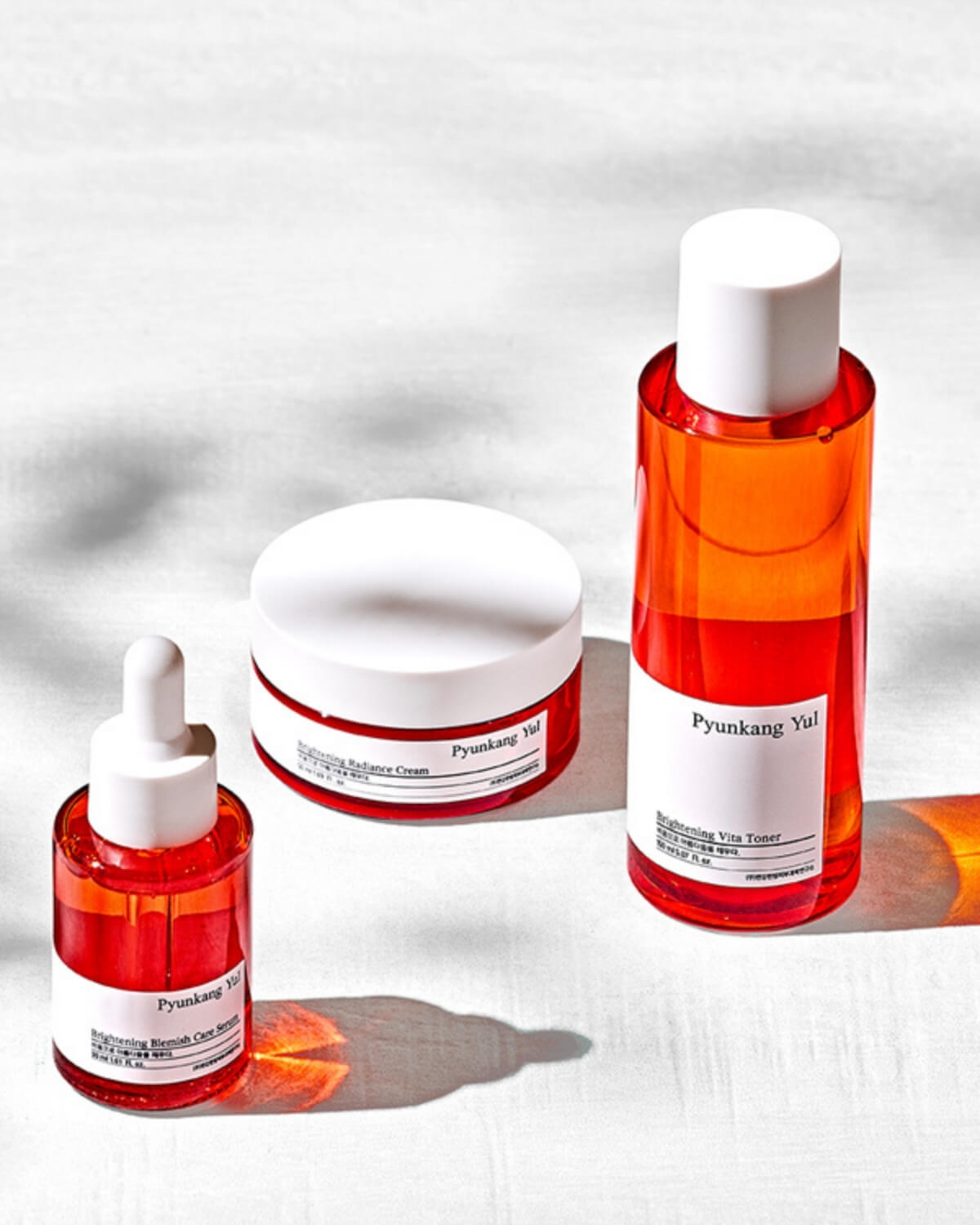 The Pyunkang Yul Brightening products (Toner, Serum, and Cream) arranged on a white, textured surface with bright lighting casting sharp shadows.