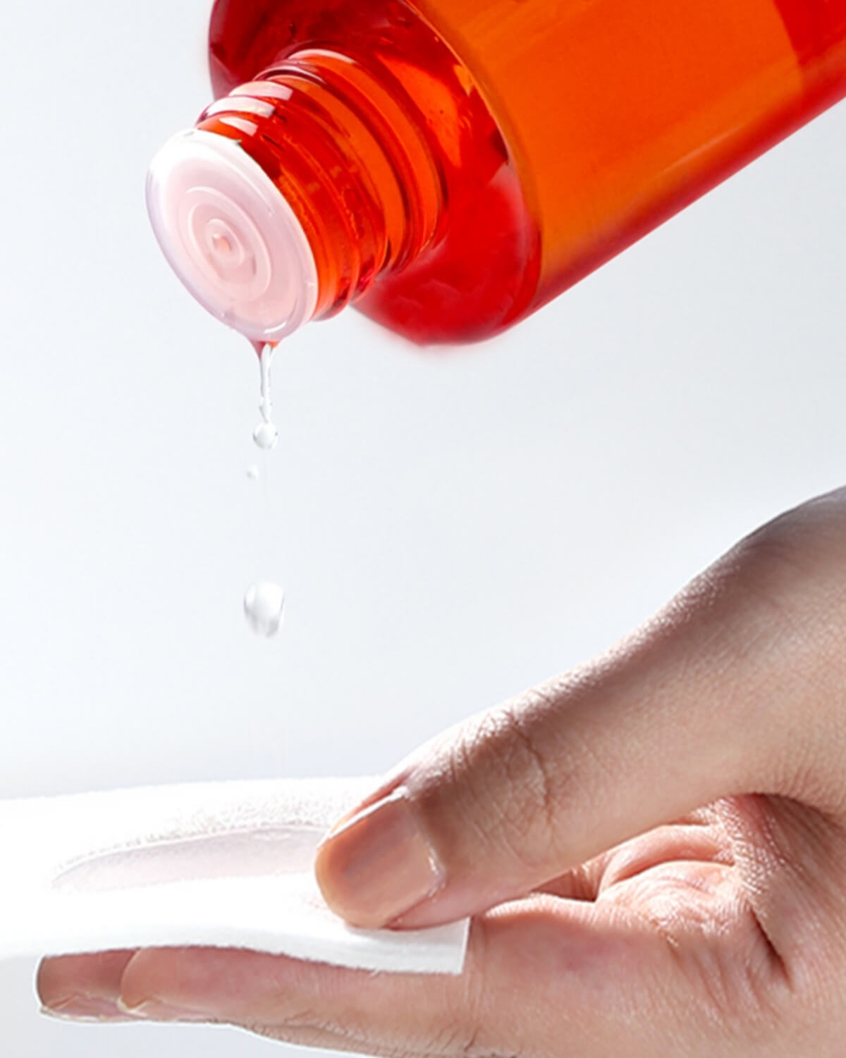 A close-up of a hand holding a white cotton pad, with drops of the orange-red Pyunkang Yul Brightening Vita Toner dripping from the bottle opening onto the pad.