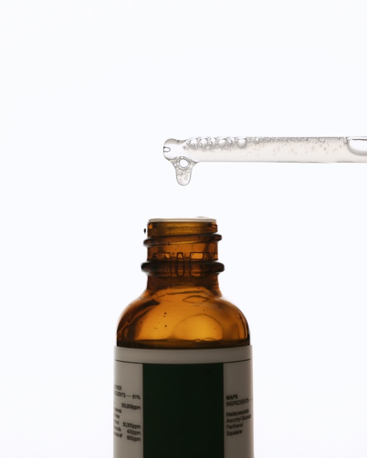 A close-up shot of an amber glass dropper bottle with its cap removed. A glass dropper is held above the opening, with a single drop of clear liquid suspended at its tip, about to fall into the bottle.