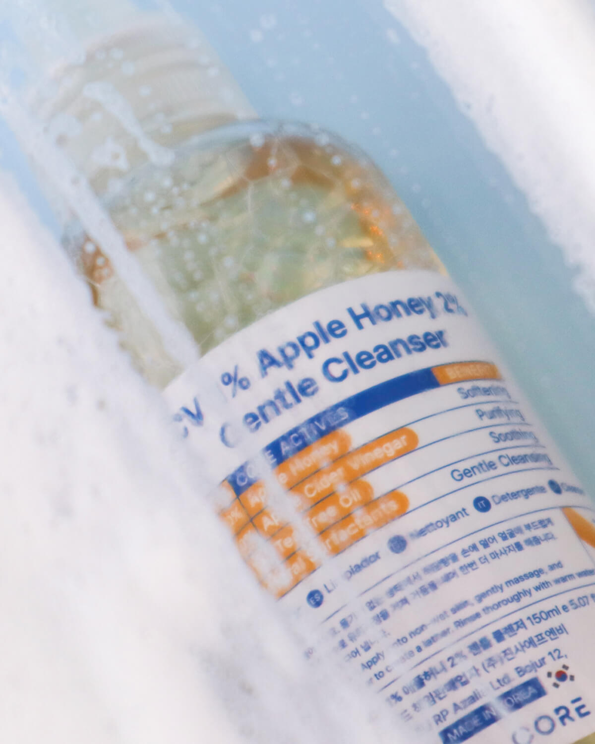 A close-up, angled view of the cleanser bottle submerged in foamy, soapy water, with the product label visible through the suds.