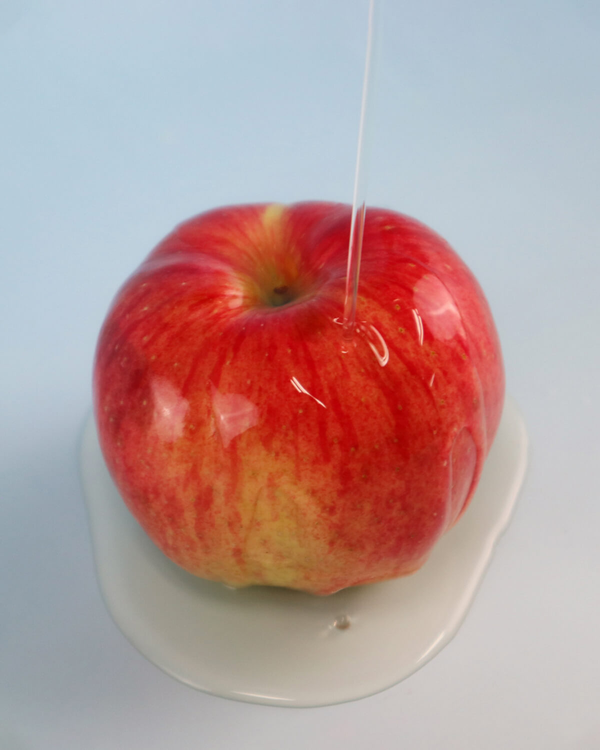 A thin stream of clear liquid, representing the cleanser, is poured onto a bright red apple resting on a white surface.