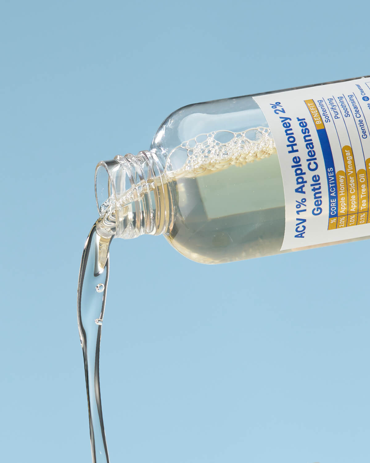 A clear, yellow liquid cleanser is being poured out of the bottle, shown at an angle against a light blue background.