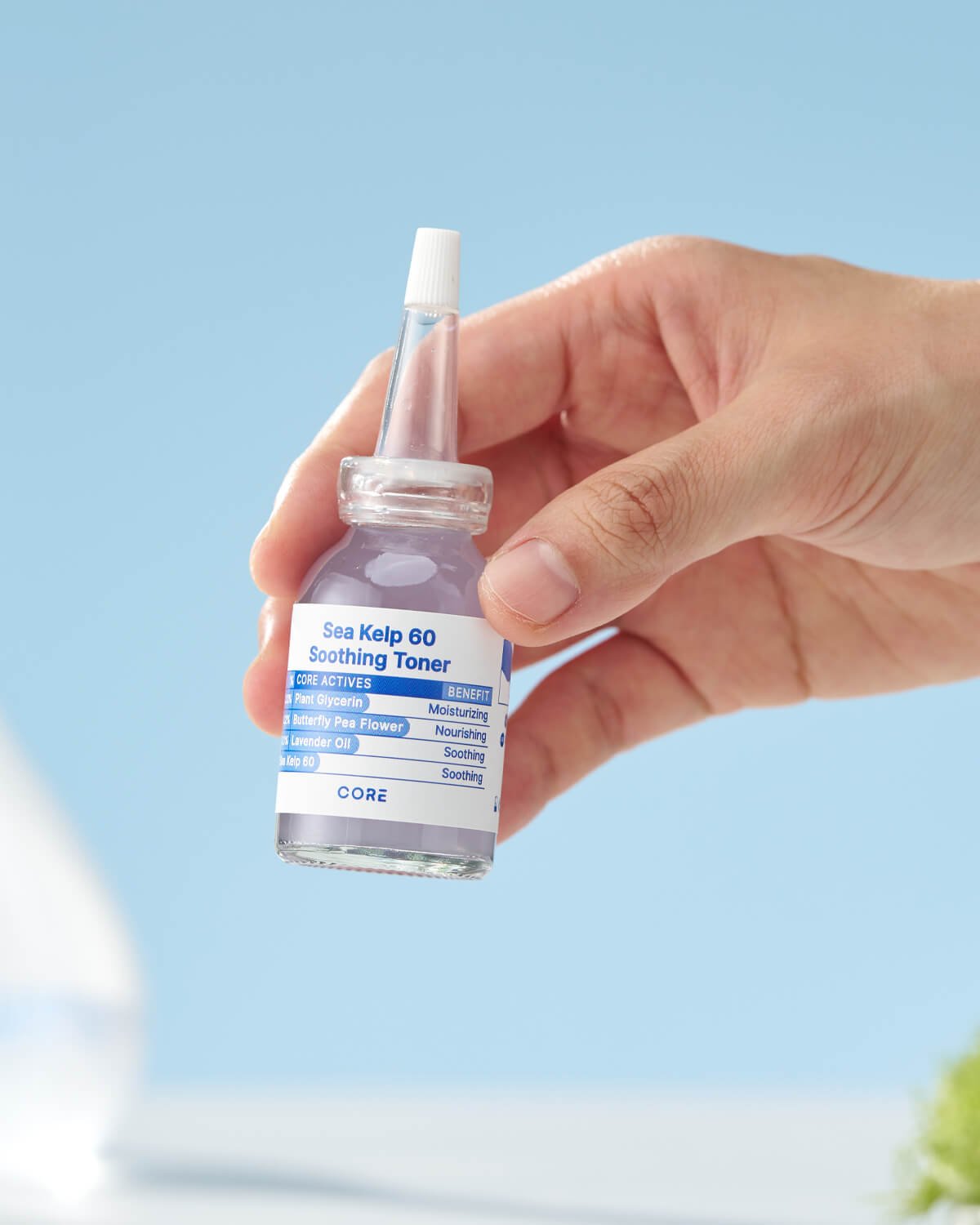 A hand holds a small sample size bottle of Core Sea Kelp 60 Soothing Toner against a bright blue background.