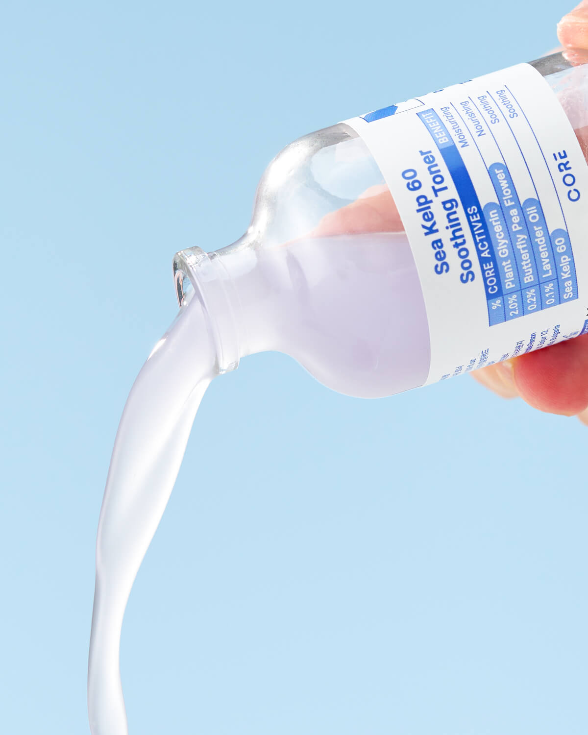 A close-up shot of the lavender-colored toner being poured out of the bottle in a thick stream against a light blue background. The label is visible.