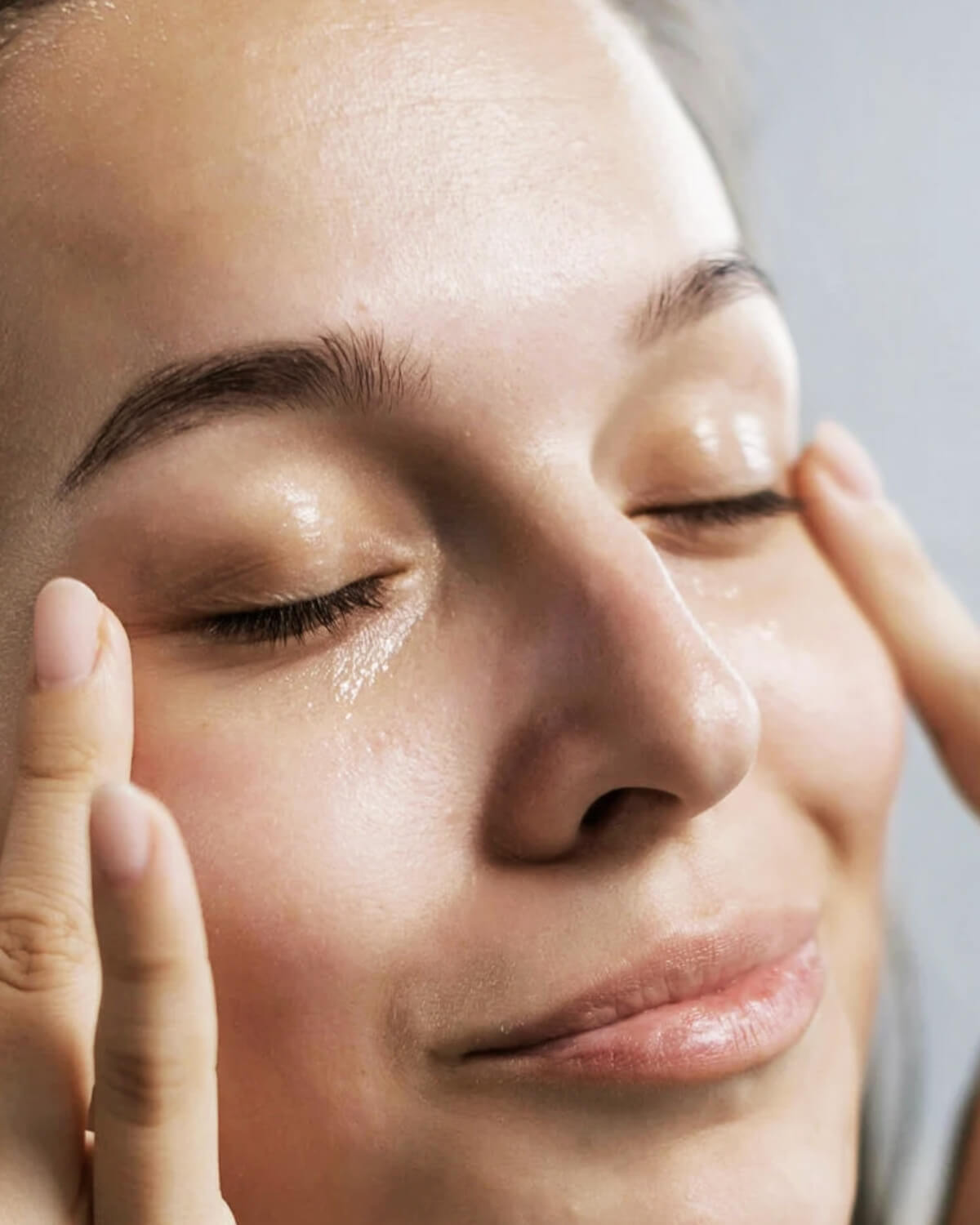 A close-up of a person's face with eyes closed, gently massaging the Dexynol-16.3 Booster Essence onto their skin, which appears dewy and hydrated.