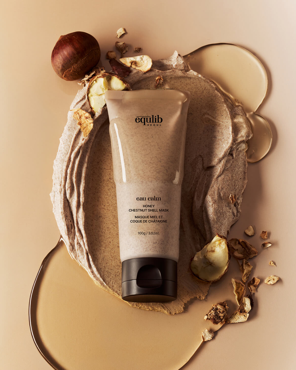 Overhead view of an Equlib Honey Chestnut Shell Mask tube resting on swirled clay texture, honey, and broken shells.