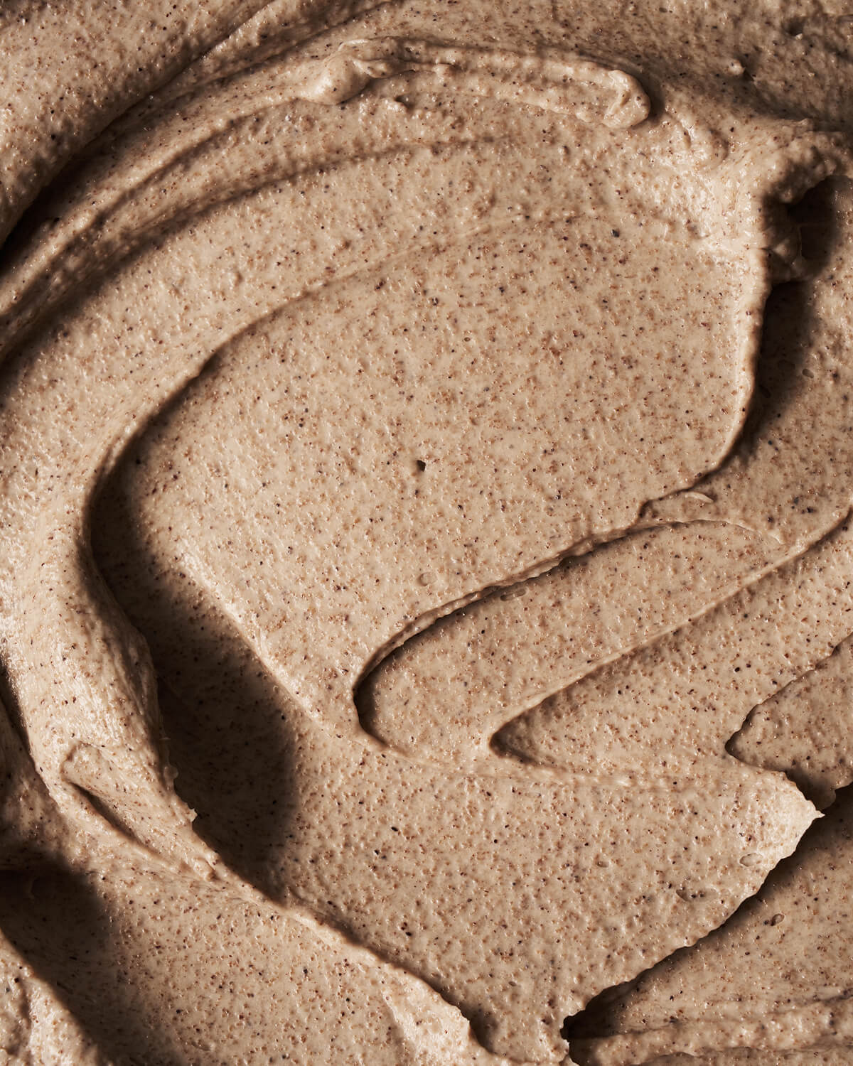 Macro texture shot of grainy, brown clay exfoliating skin mask showing ridges and small exfoliating particles.
