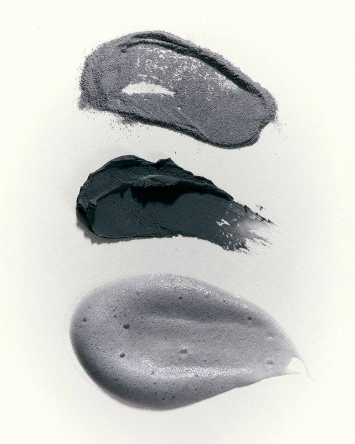 Swatches of grey exfoliating charcoal powder, dark grey face mask paste, and foamy facial cleanser lather on white surface.
