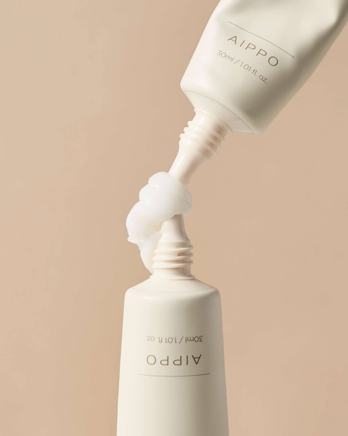 An artistic, close-up shot showing cream being squeezed directly from the nozzle of one tube to the open nozzle of another tube against a pale background. The white cream forms a soft, knot-like shape between the two tubes.
