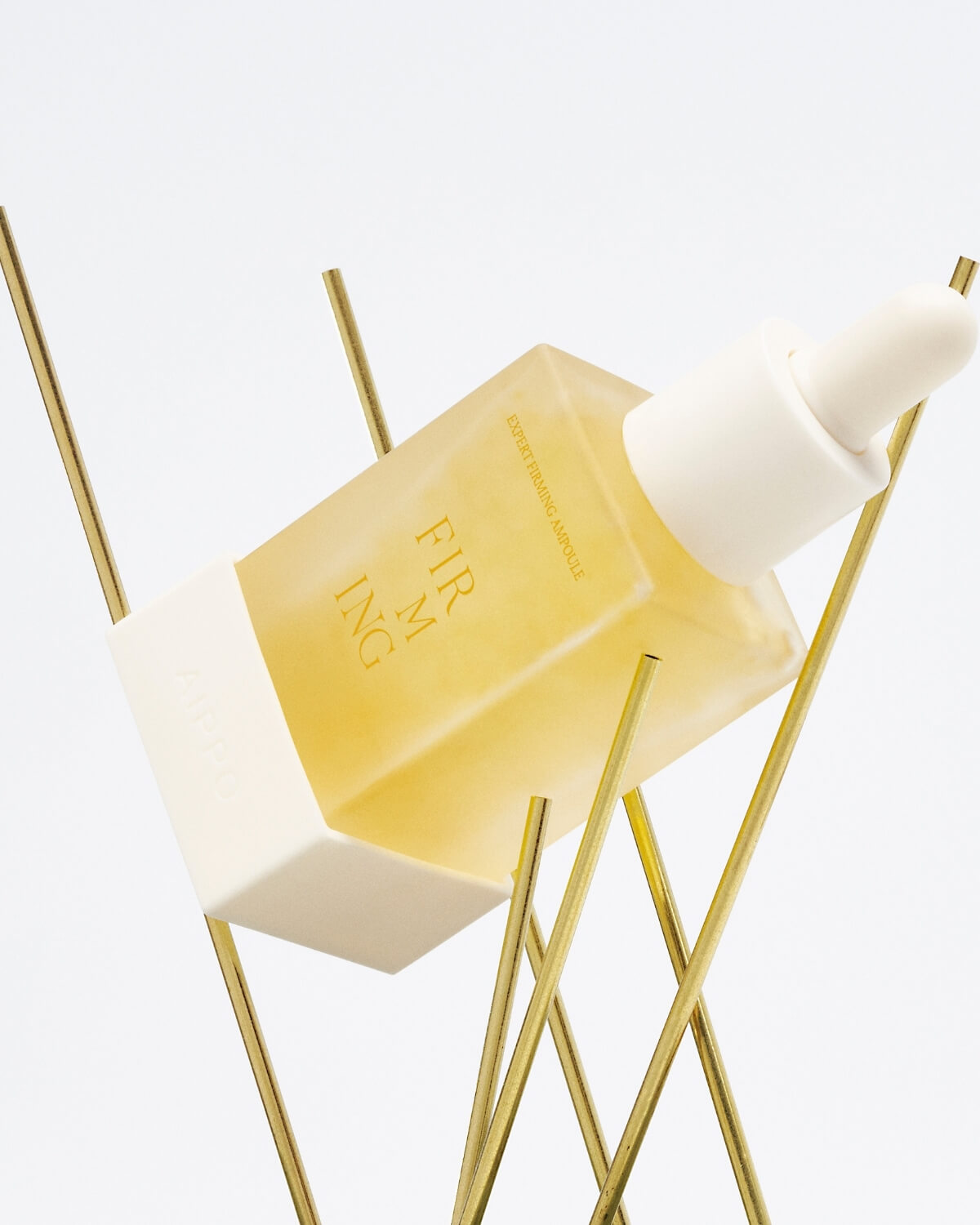 A bottle of AIPPO Expert Firming Ampoule, with a yellow-tinted liquid and visible bubbles, held diagonally by thin, crisscrossing gold metal rods against a white background. The text "EXPERT FIRMING AMPOULE," "FIRMING," and "AIPPO" is visible on the bottle.