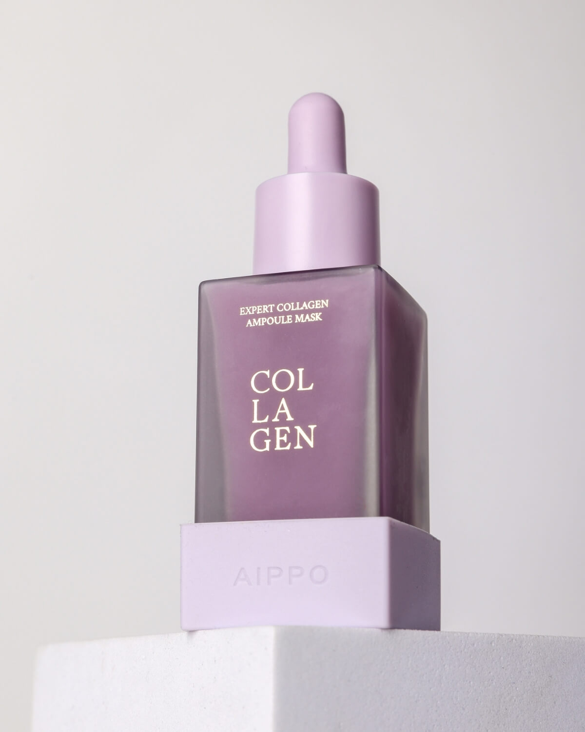 A close-up studio shot of the purple Aippo Collagen Ampoule Mask bottle sitting on a white geometric platform, dramatically lit to showcase the velvety, frosted texture of the glass.