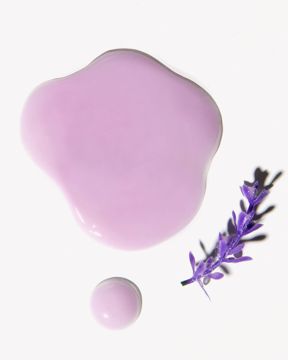 An aerial view of a smooth, glossy puddle of the light purple ampoule mask product, next to a small, rounded drop and a sprig of purple lavender, on a white surface.