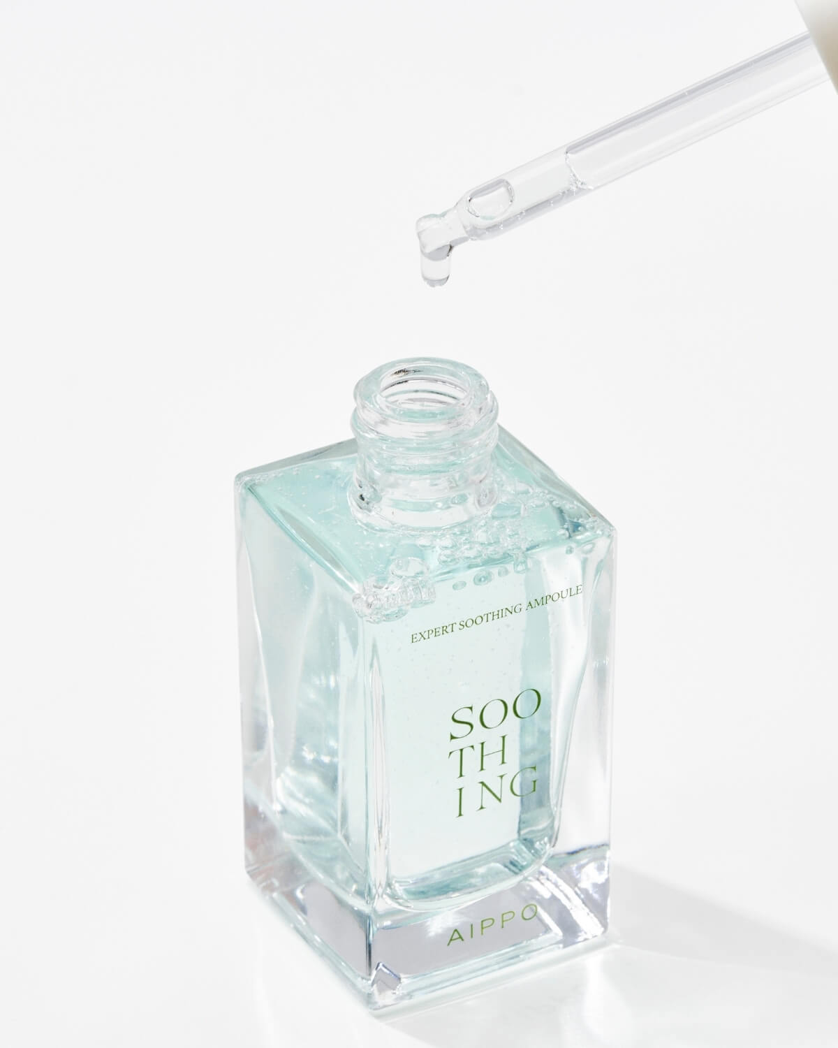 A close-up of the open bottle of AIPPO Expert Soothing Ampoule. A clear glass dropper is held above the neck, with a single drop of the light blue liquid product dripping back into the bottle. The text "EXPERT SOOTHING AMPOULE" is visible on the side.