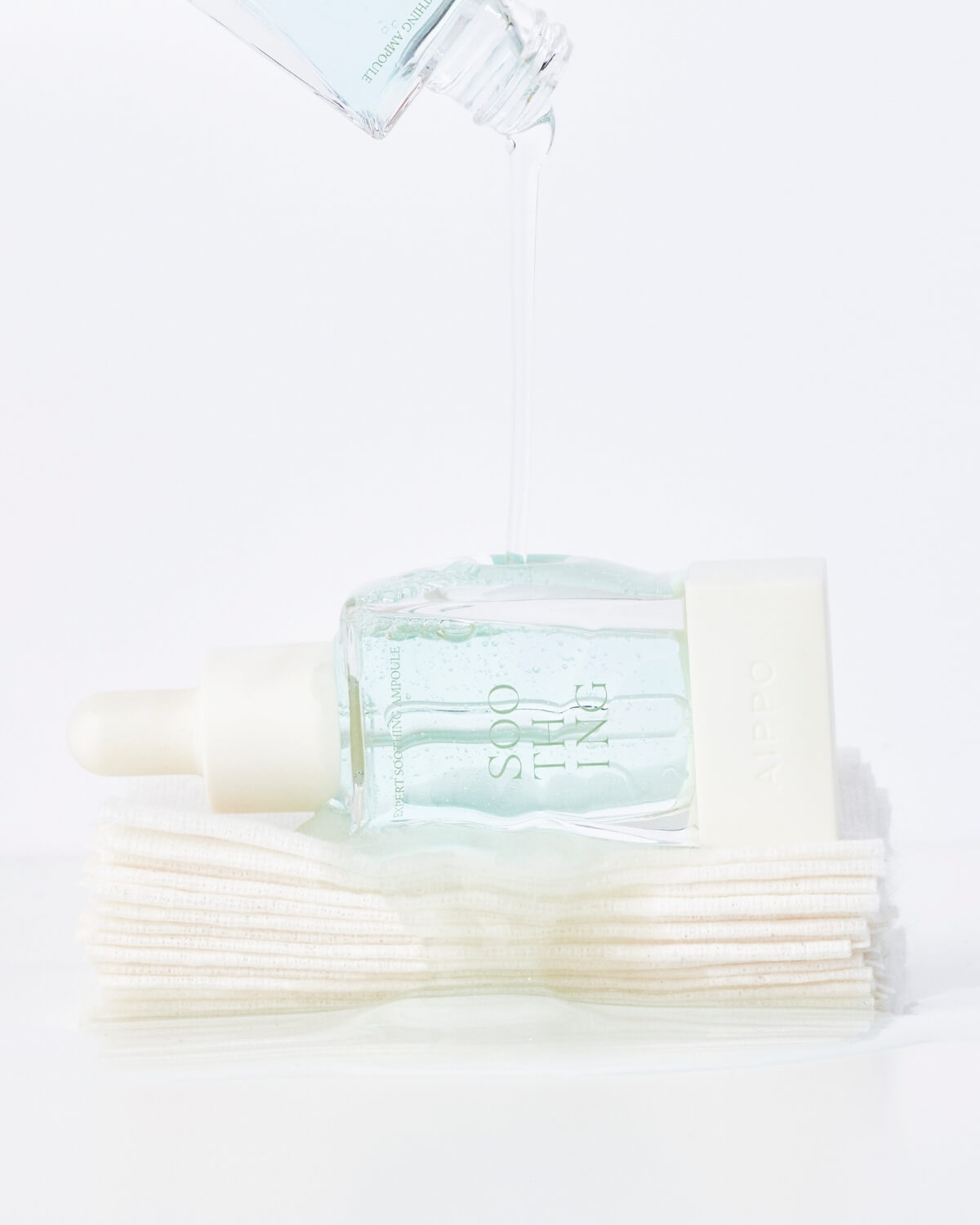 A creative product shot where the AIPPO Expert Soothing Ampoule bottle is lying on its side on a stack of white cotton pads. A thin stream of the clear, light blue liquid is being poured onto the bottle and pads from another source above.