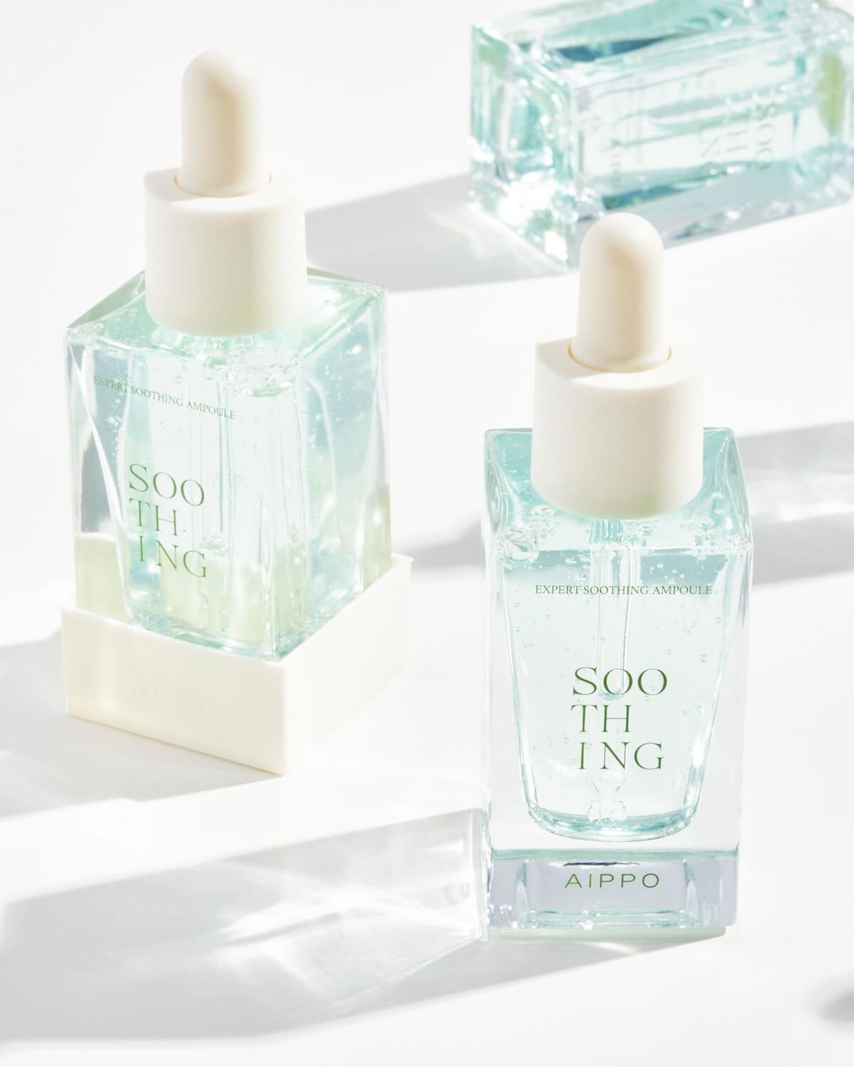 Two bottles of AIPPO Expert Soothing Ampoule are positioned on a white surface, casting long shadows. The bottle in the foreground is clearly labeled, and a third, partially visible bottle lies out of focus in the upper right corner.