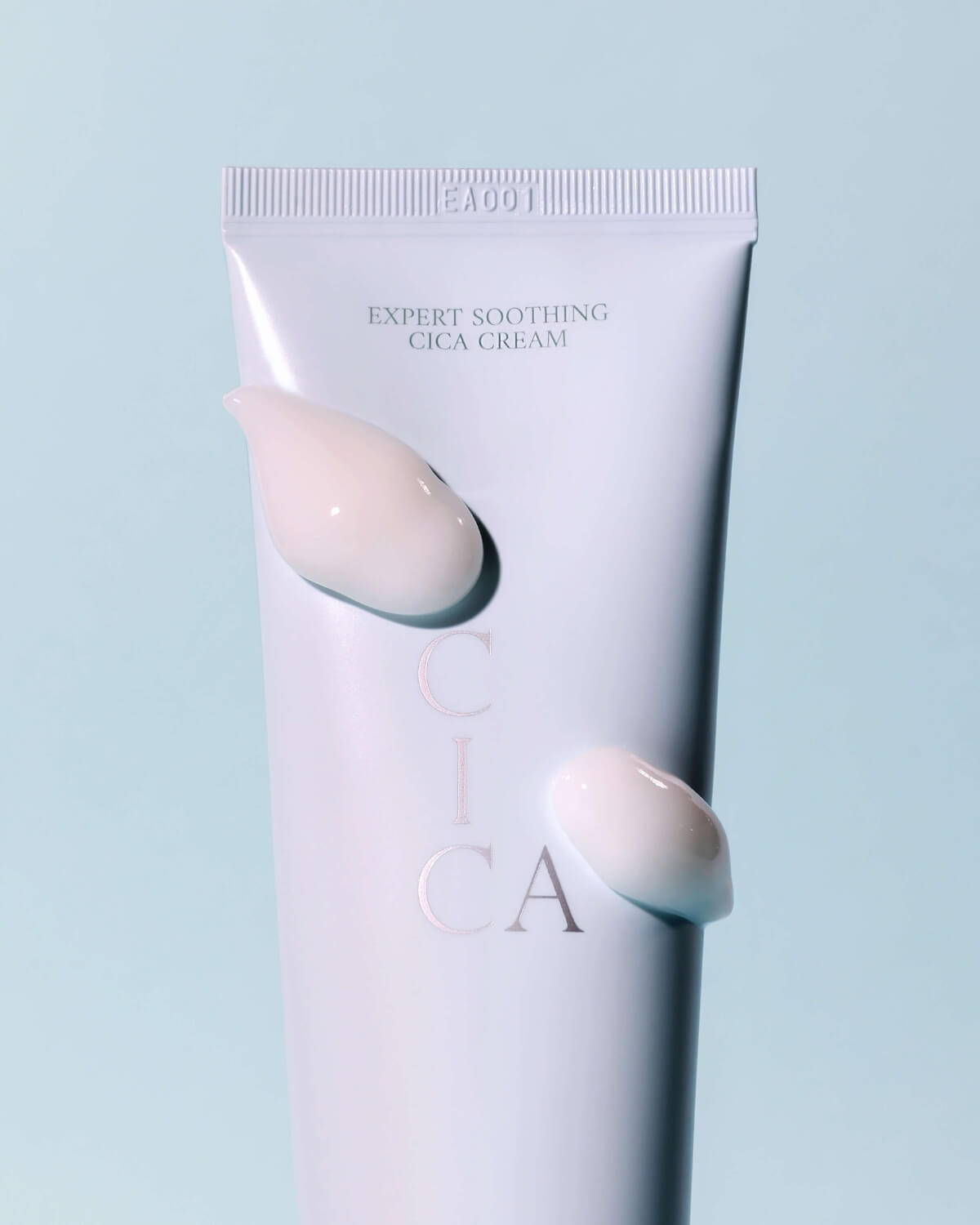 A close-up of the Aippo Expert Soothing Cica Cream tube against a soft blue background, with two smooth dollops of the white cream product resting on its surface.