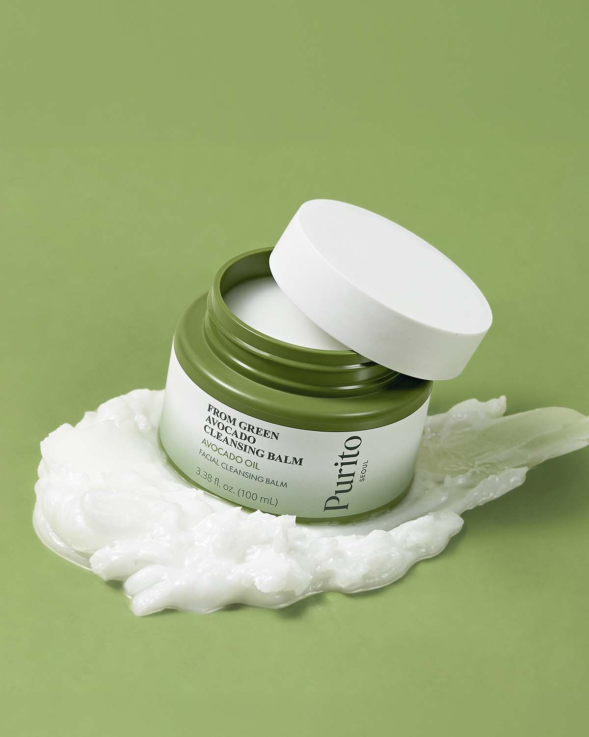 Purito Avocado Cleansing Balm jar tilted on a swirl of creamy white facial cleanser over a green surface.
