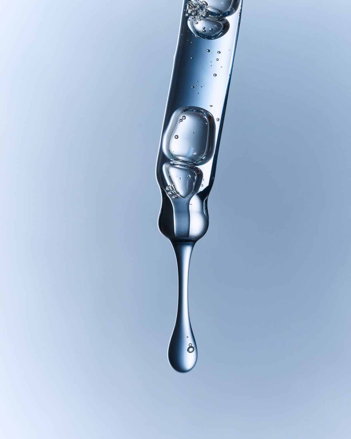 Close-up of a glass dropper releasing a clear skincare liquid droplet against a blue background.