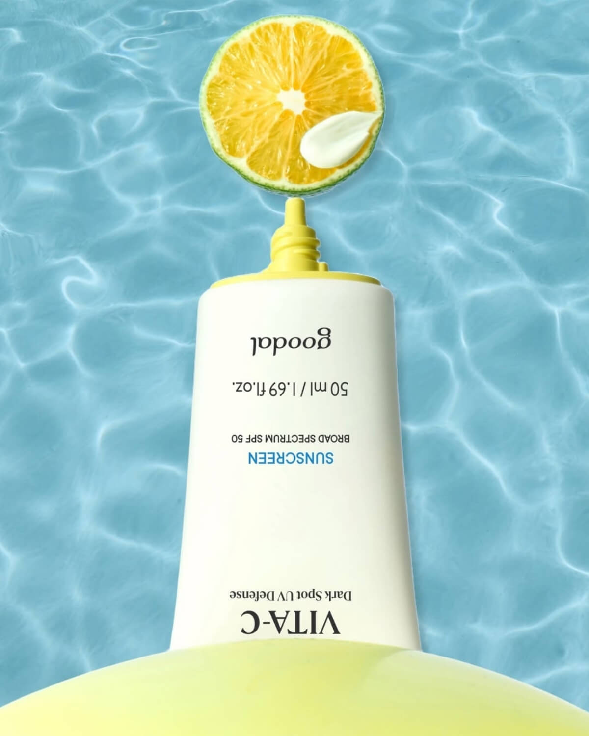 Goodal green tangerine sunscreen tube pointing down toward a fresh slice of citrus fruit over clear, rippling blue water.