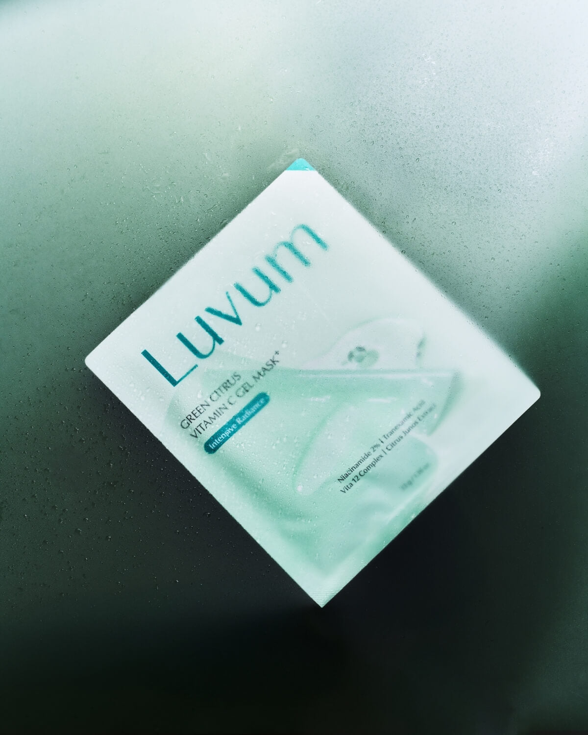 A wet Luvum Green Citrus Vitamin C Gel Mask packet resting at an angle behind a glass pane covered in mist.