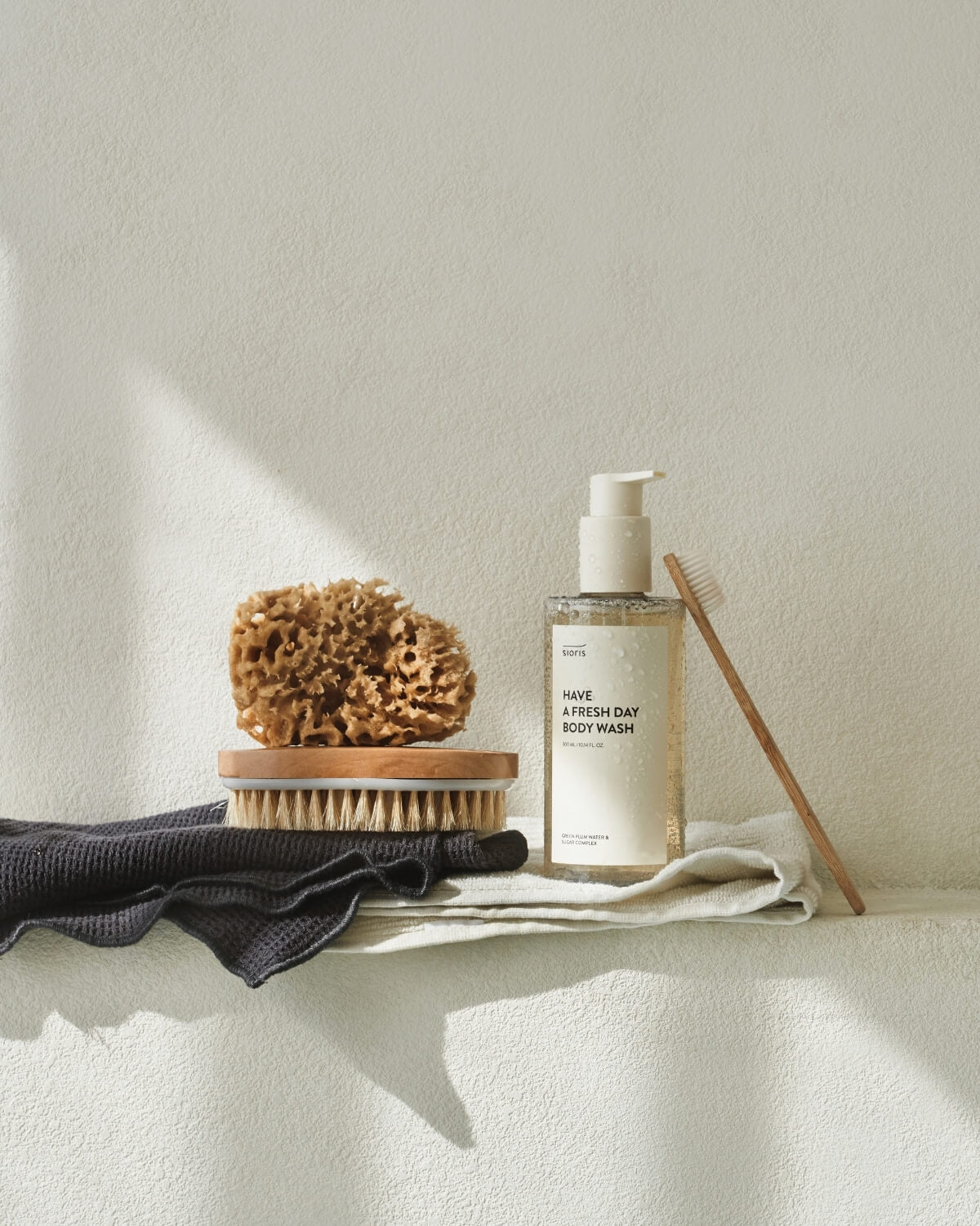 Sioris Have a Fresh Day Body Wash bottle on a towel next to a natural sea sponge, bath brush, and toothbrush.