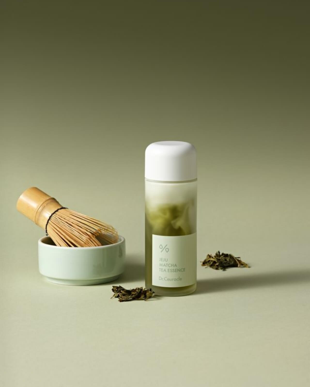 Jeju Matcha Tea Essence bottle next to a traditional bamboo matcha whisk and dried green tea leaves.