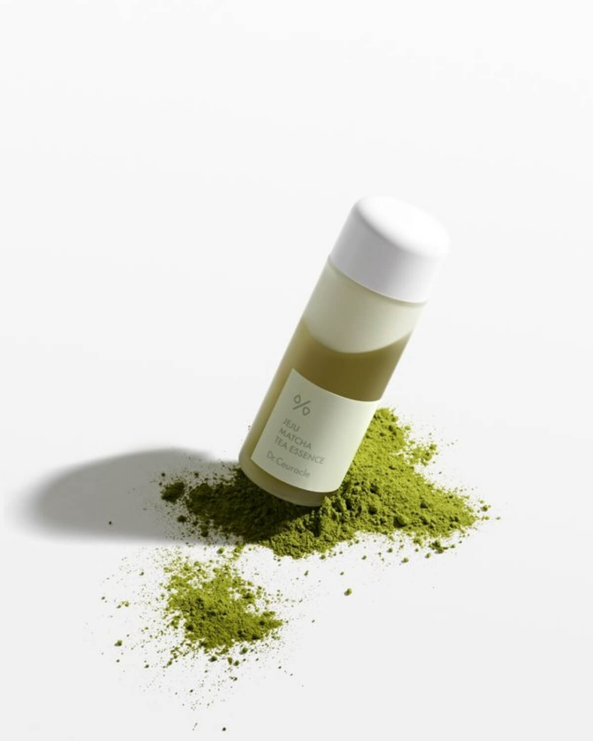 A bottle of Dr. Ceuracle Jeju Matcha Tea Essence leaning against a pile of vibrant green matcha powder.
