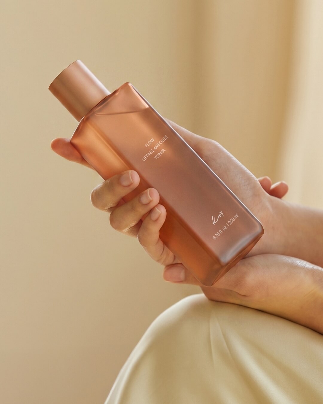 A person holding a 200ml bottle of Koy Flow Lifting Ampoule Toner in both hands against a warm beige background.