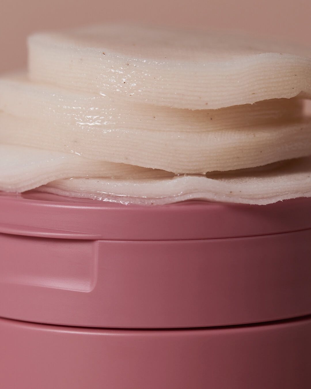 Detailed close-up of a stack of wet cotton pads resting on top of a pink skincare container.