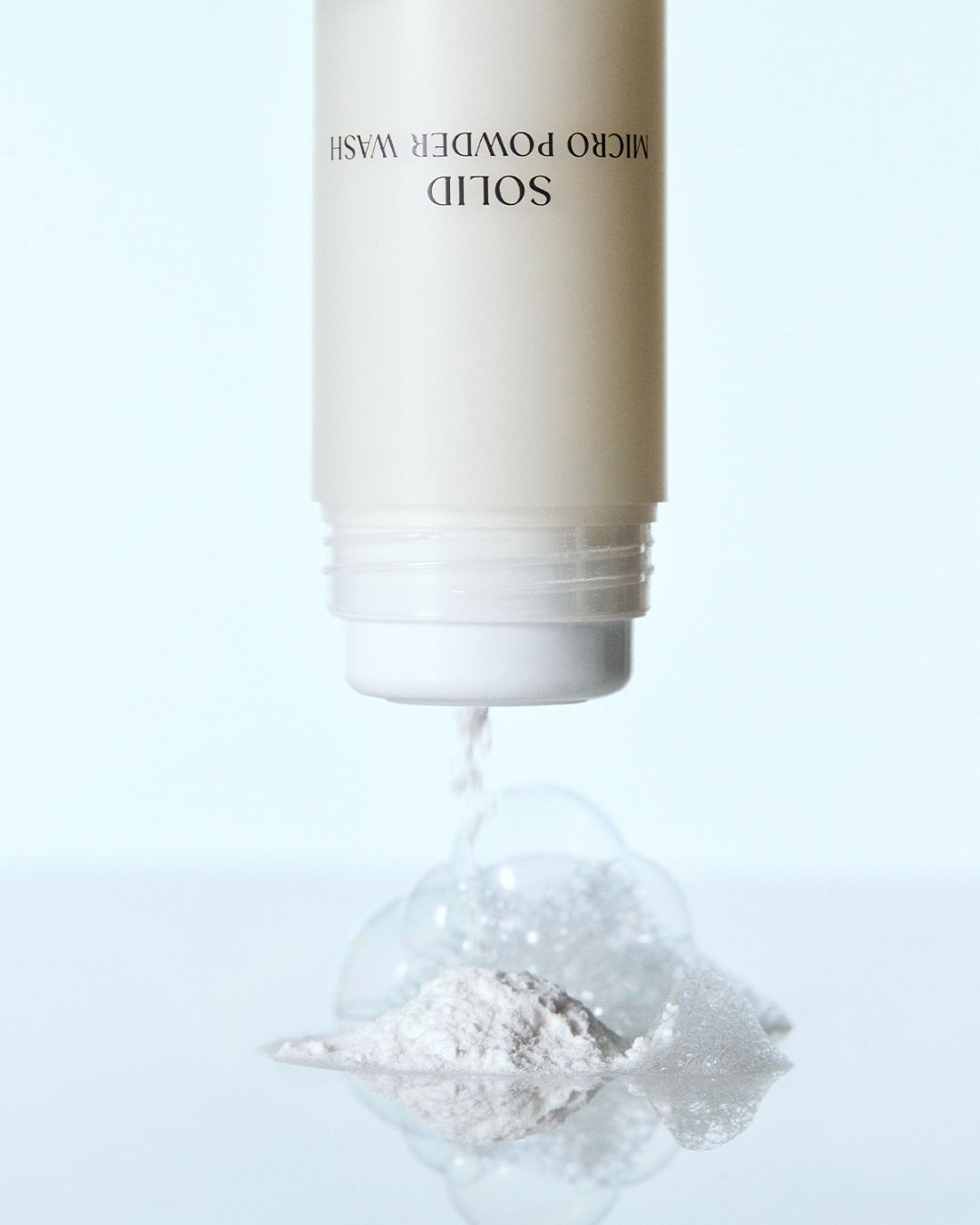 White exfoliating enzyme powder pouring from a Koy Solid Micro Powder Wash bottle onto a soapy surface.