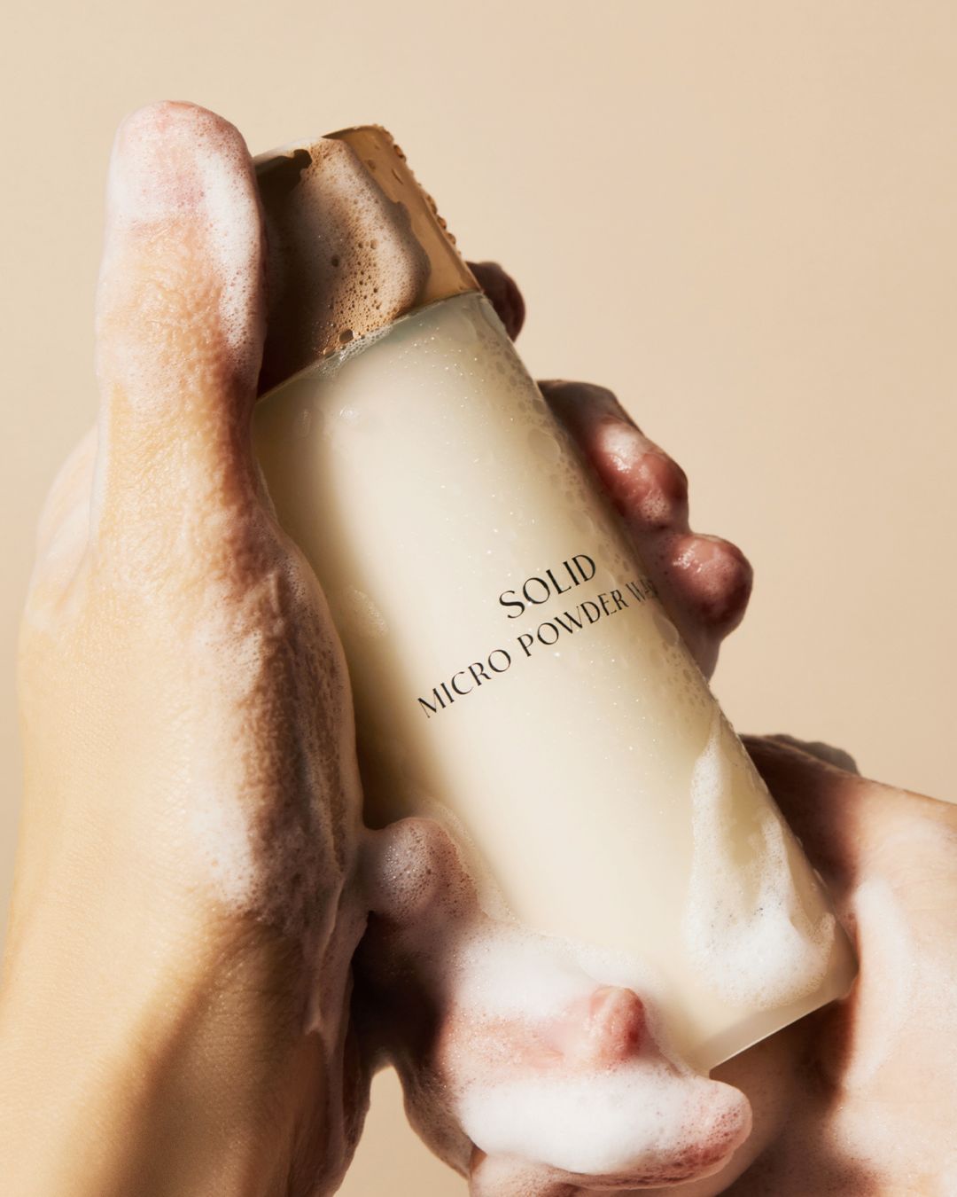 Foamy soapy hands holding a bottle of Koy Solid Micro Powder Wash facial cleanser against a beige background.