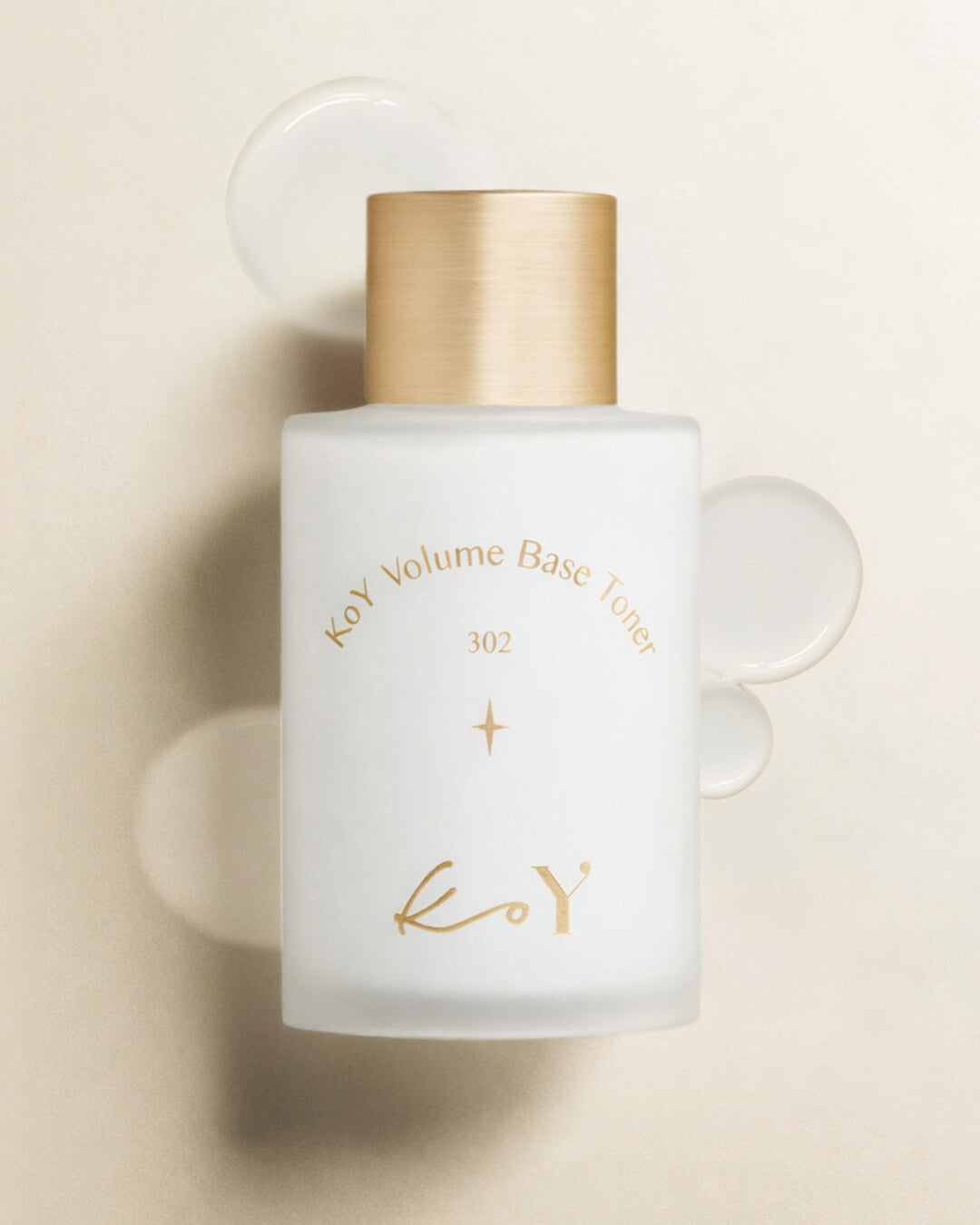 Flat lay of a KoY Volume Base Toner bottle surrounded by clear drops of liquid gel texture.