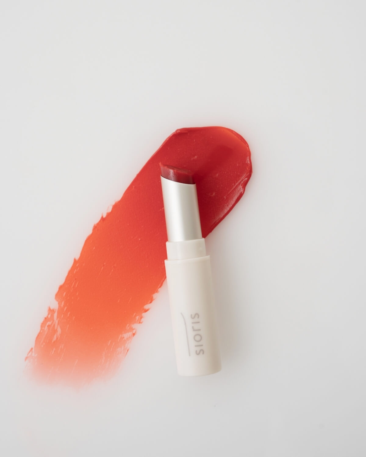 Sioris LipSafer Color Balm in Reddish positioned vertically against a clean, swiped red-orange lip tint smudge.