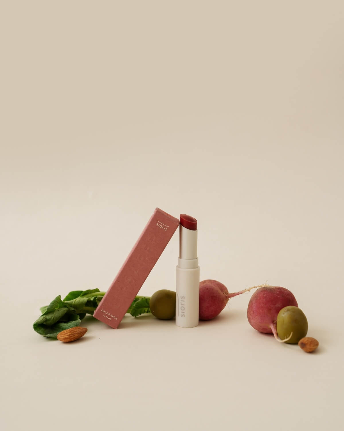 Sioris LipSafer Color Balm Reddish tube with its pink box, styled with fresh radishes, olives, and almonds.