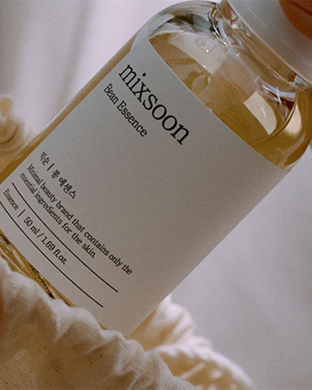 An extreme close-up of the Mixsoon Bean Essence clear glass bottle, angled to show the minimalist white label with the text 'Bean Essence' and the volume '50 ml / 1.69 fl oz,' emphasizing the light yellow-gold color of the essence against a soft, white textile background.