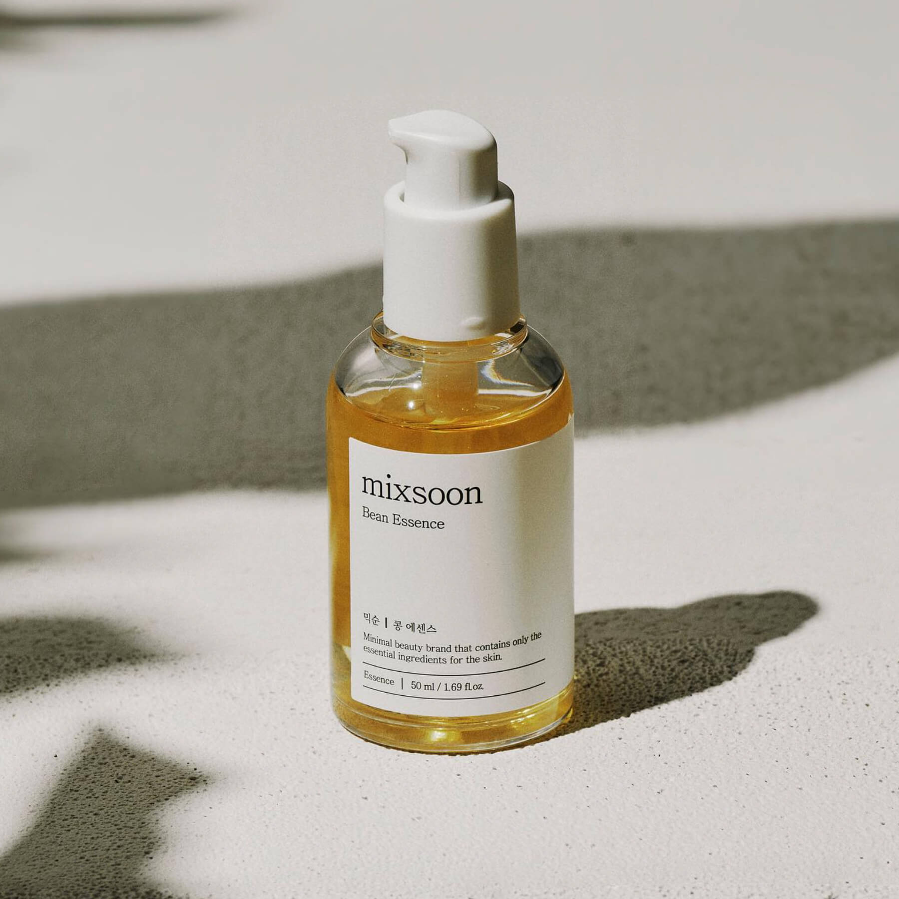 A bright, sunlit studio shot of the Mixsoon Bean Essence bottle, showcasing the light yellow-gold essence inside. The bottle is set on a light surface with a strong, diagonal shadow cast across the background, emphasizing its clean, minimalist design.