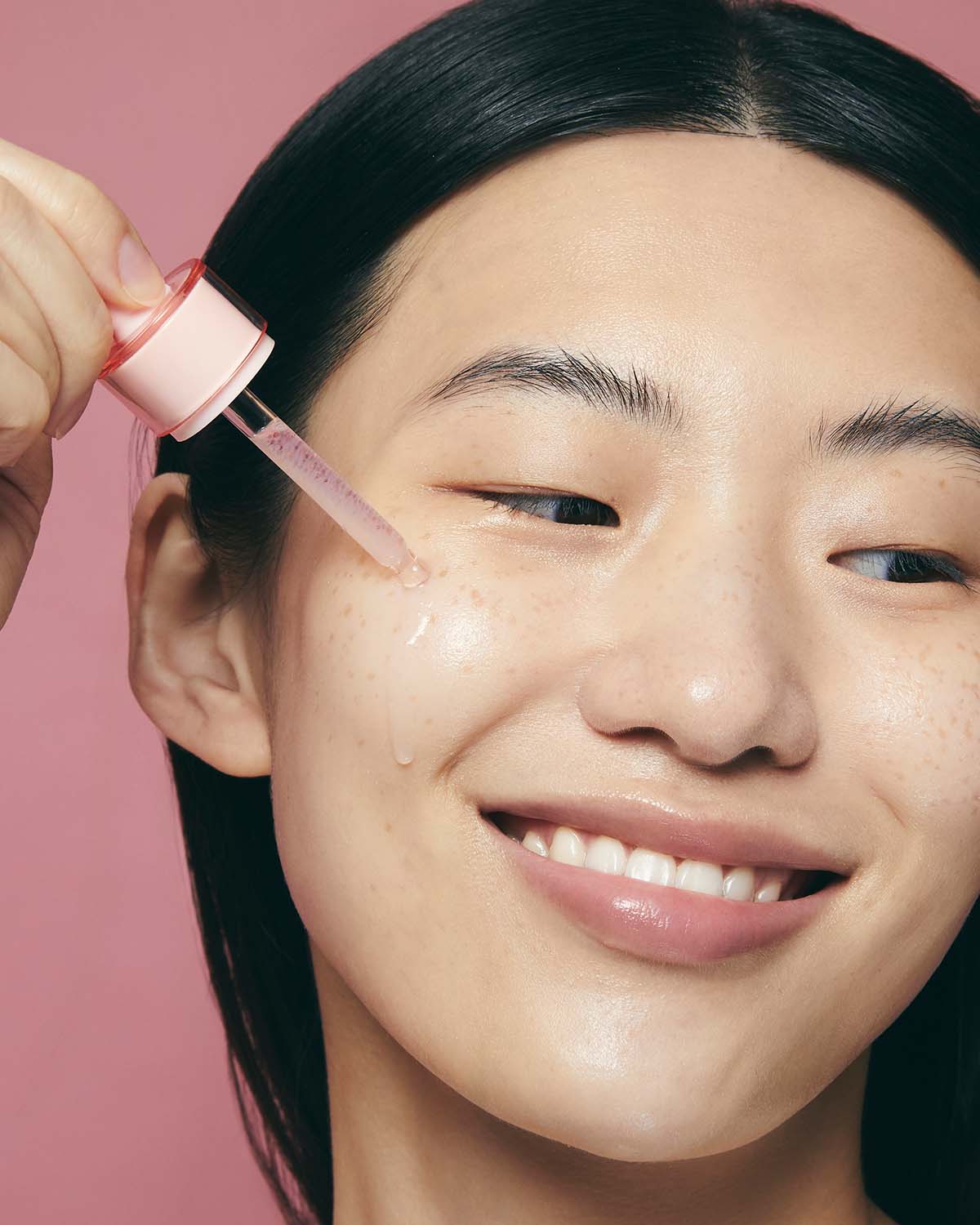 A smiling woman applying a drop of Purito Multi PDRN Collagen EGF Serum to her cheek with a dropper.