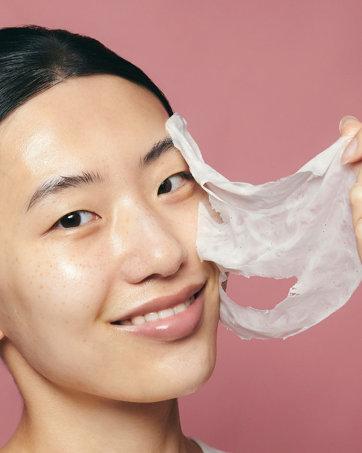 A smiling woman peeling off a dried white Purito Multi PDRN Collagen Real Glow Mask from her face.
