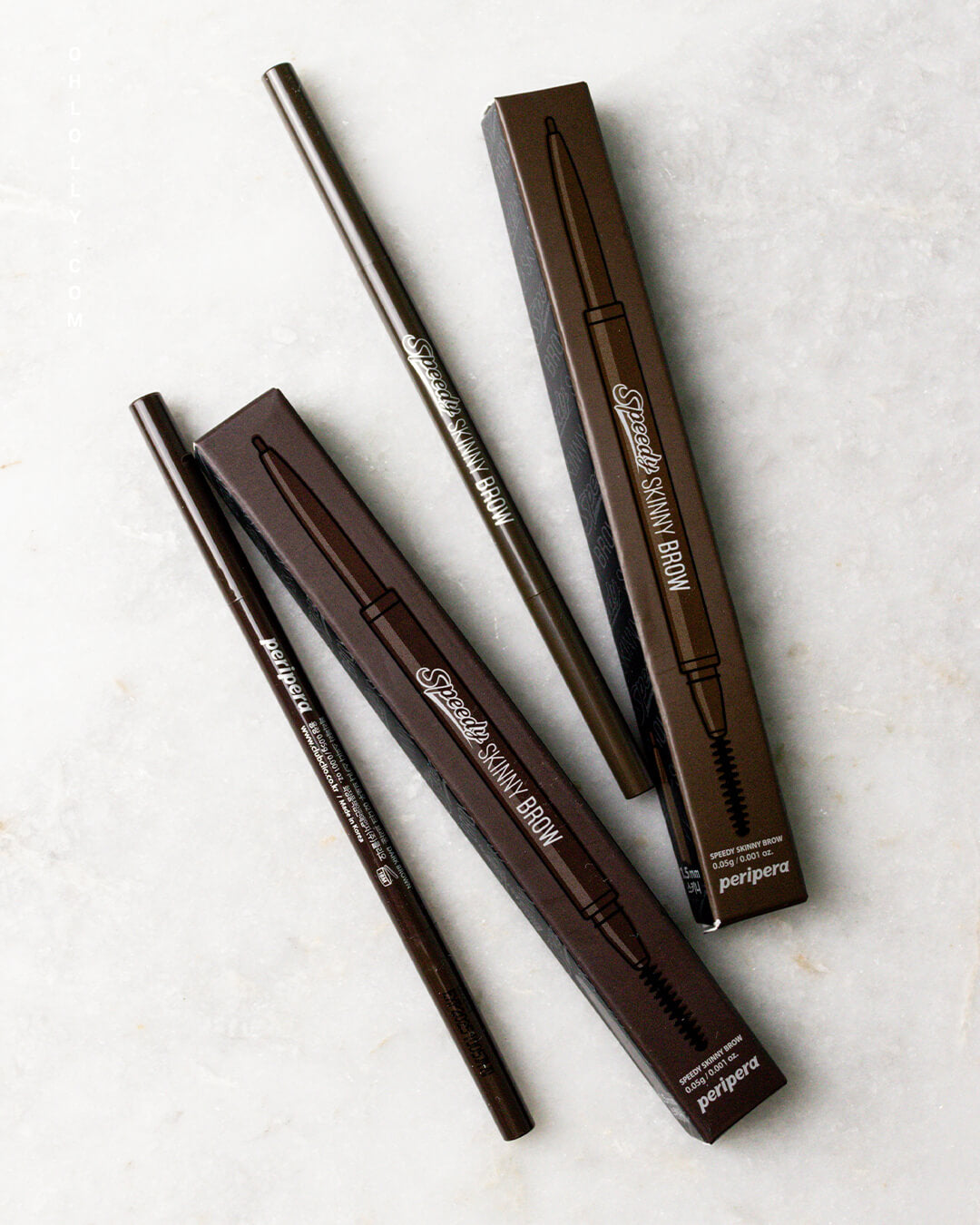 Two Peripera Speedy Skinny Brow pencils and their dark brown cardboard boxes are arranged diagonally on a white marble-like surface, showcasing the ultra-slim 1.5mm pencil design and the 'SPEEDY SKINNY BROW' product name.