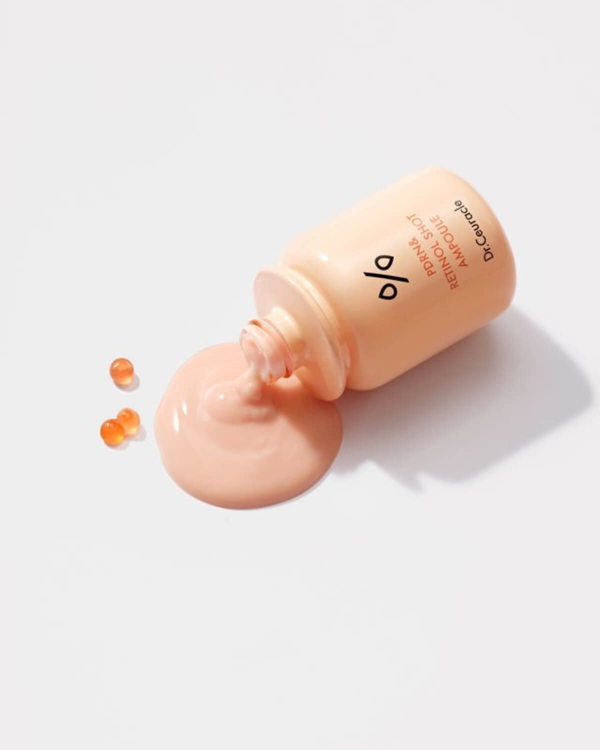 PDRN & Retinol Shot Ampoule spilled on a white surface with small orange active ingredient beads.