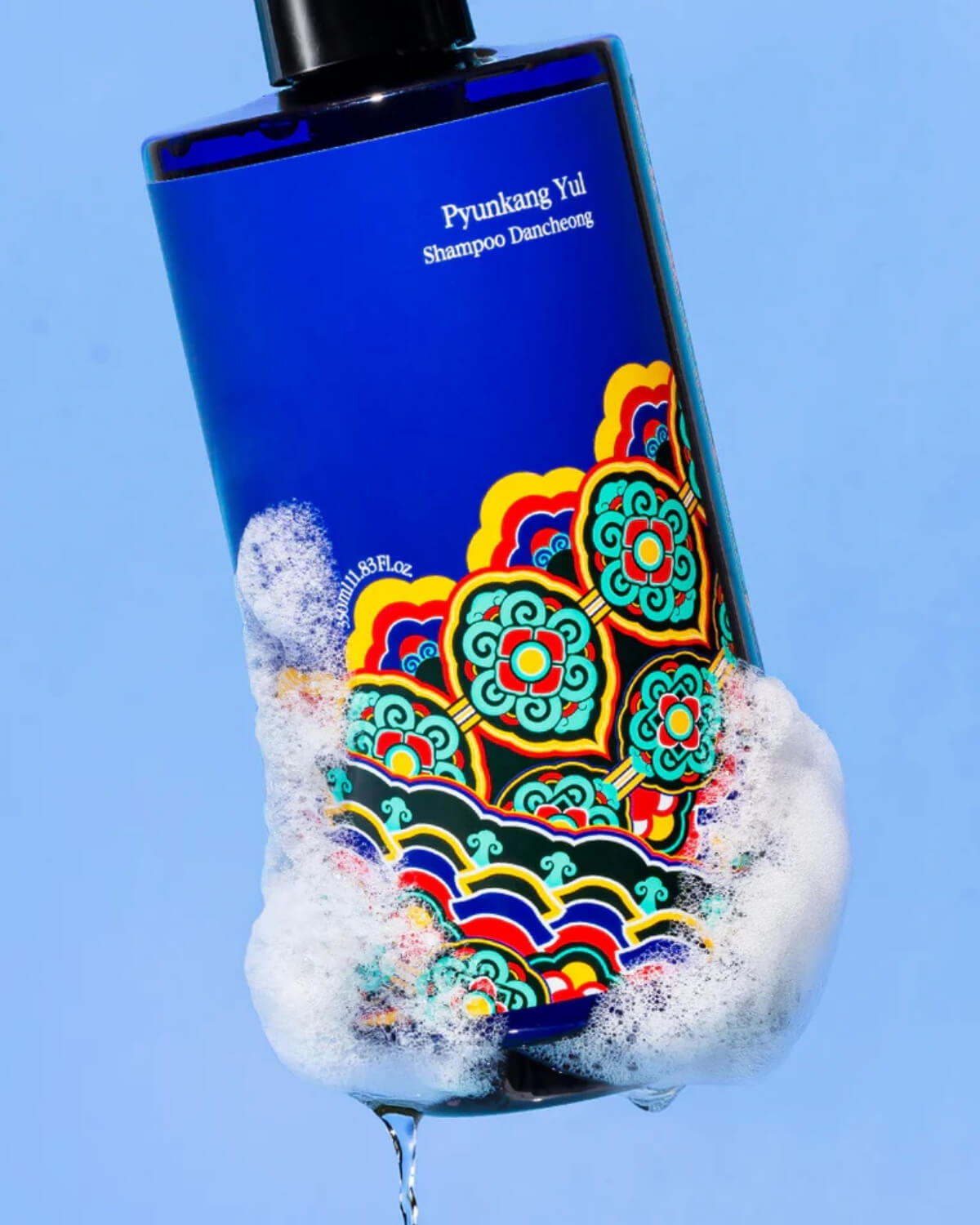 A close-up, tilted shot of the Pyunkang Yul Shampoo Dancheong bottle covered in white soap suds and bubbles against a solid light blue background.