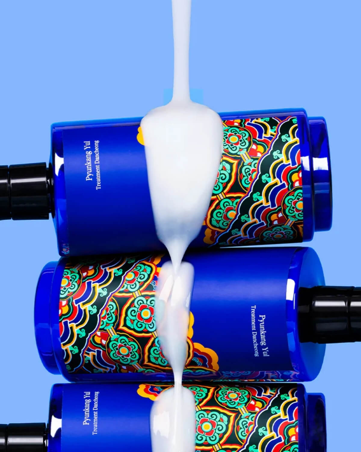Three blue treatment bottles stacked horizontally with a thick, white cream pouring over them against a bright blue background.