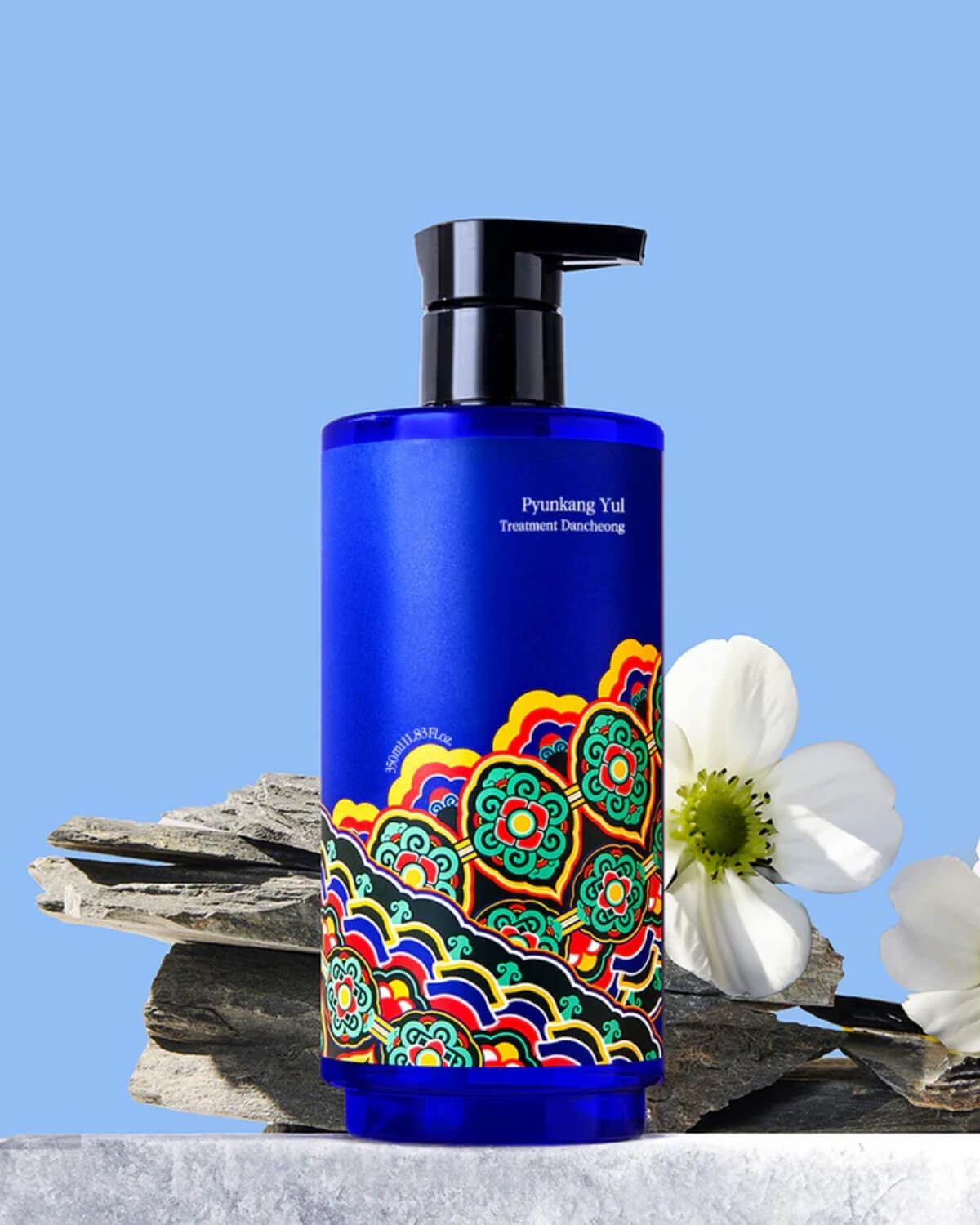 The blue treatment bottle standing on a grey rock next to a white flower, set against a solid light blue background.