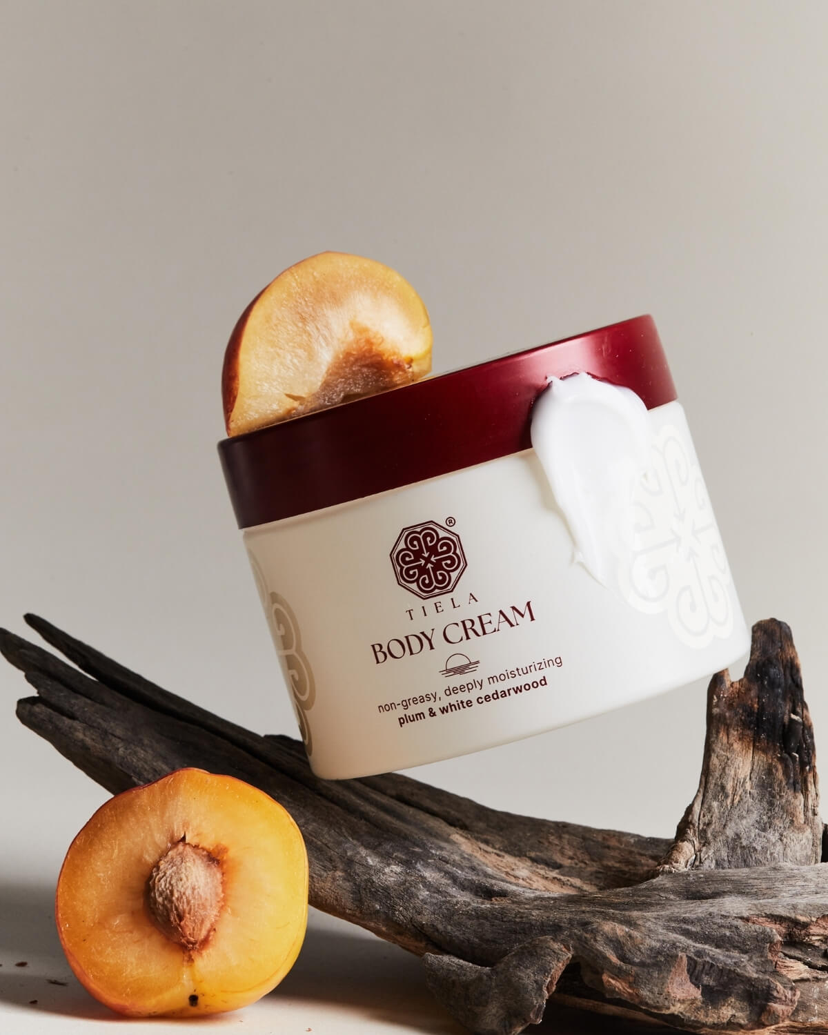 Tiela Body Cream jar tipped on its side against a piece of driftwood, with a plum slice resting on top and a whole plum half in the foreground.