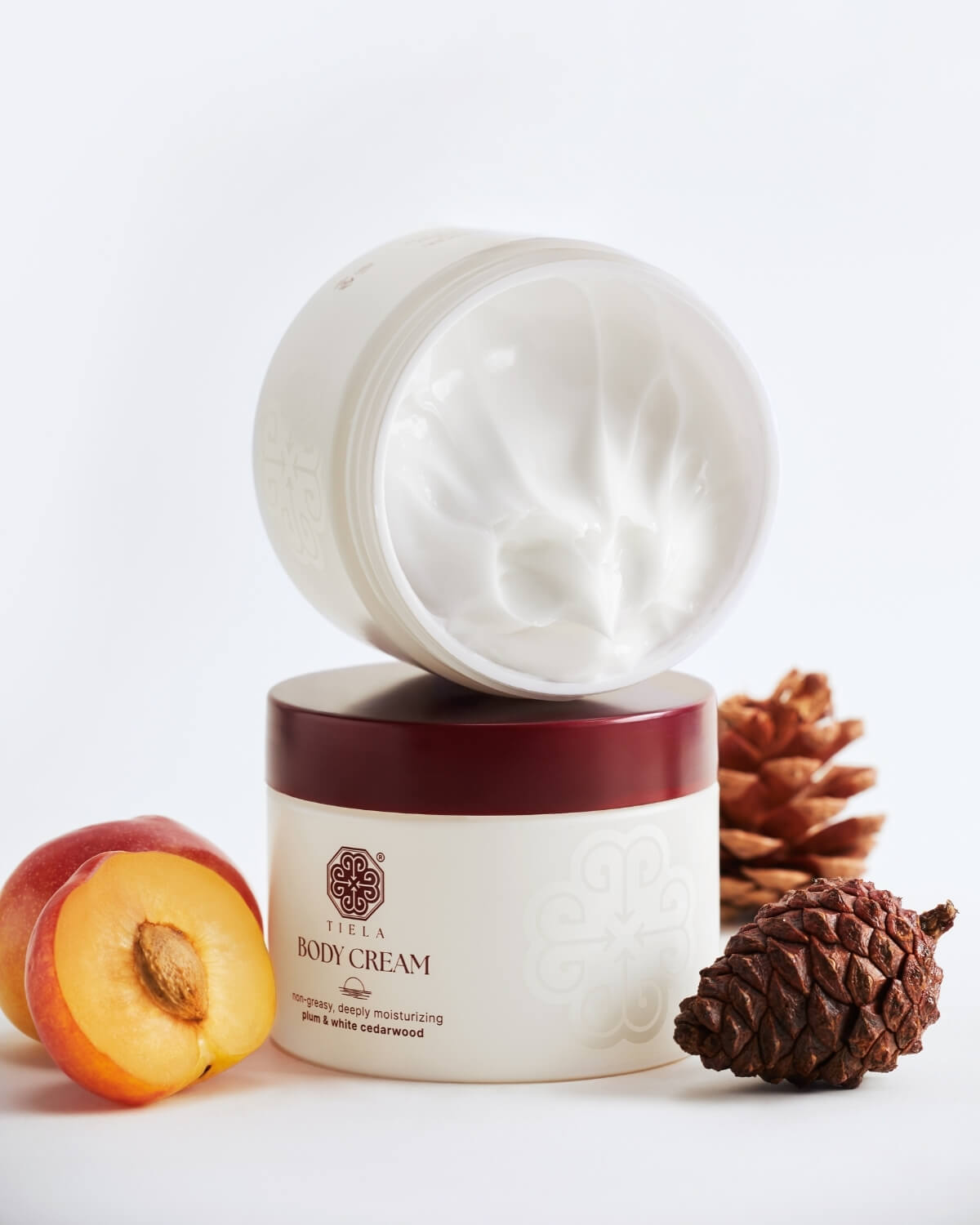 Two Tiela Body Cream jars stacked vertically, with the top jar open to show the white cream, surrounded by sliced plum and a pine cone.