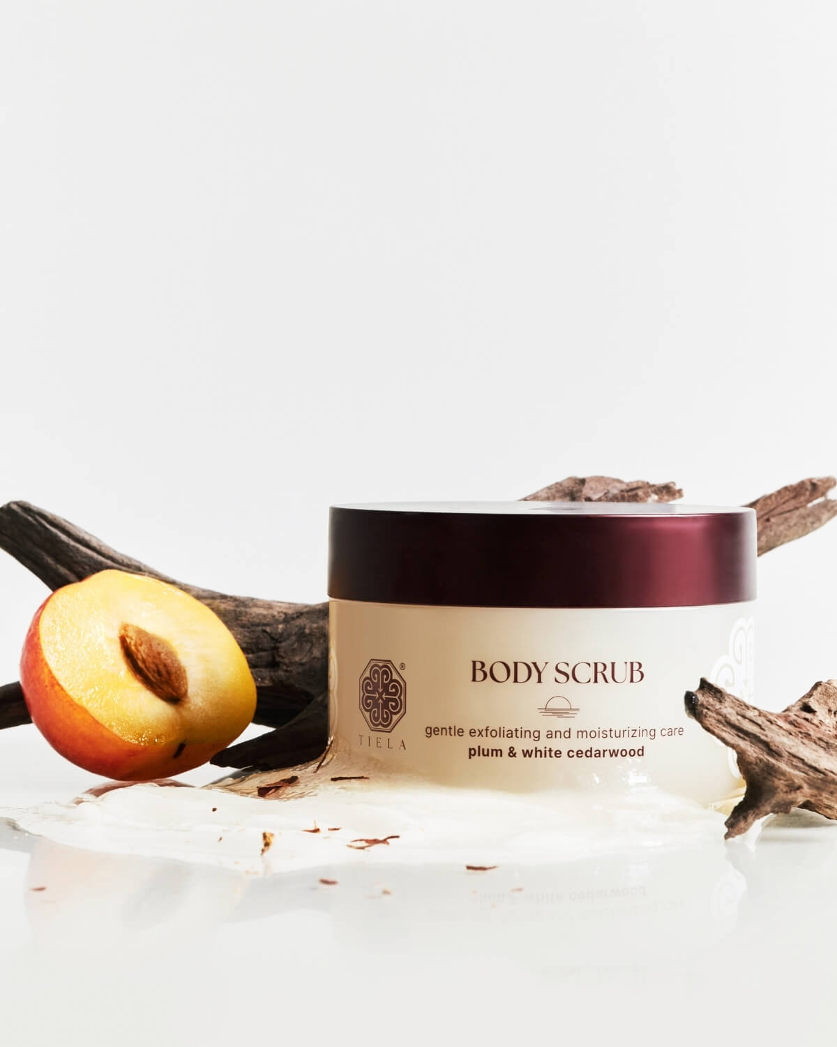 Tiela Body Scrub jar sitting on a pool of the scrub product, surrounded by dark driftwood and a halved plum.