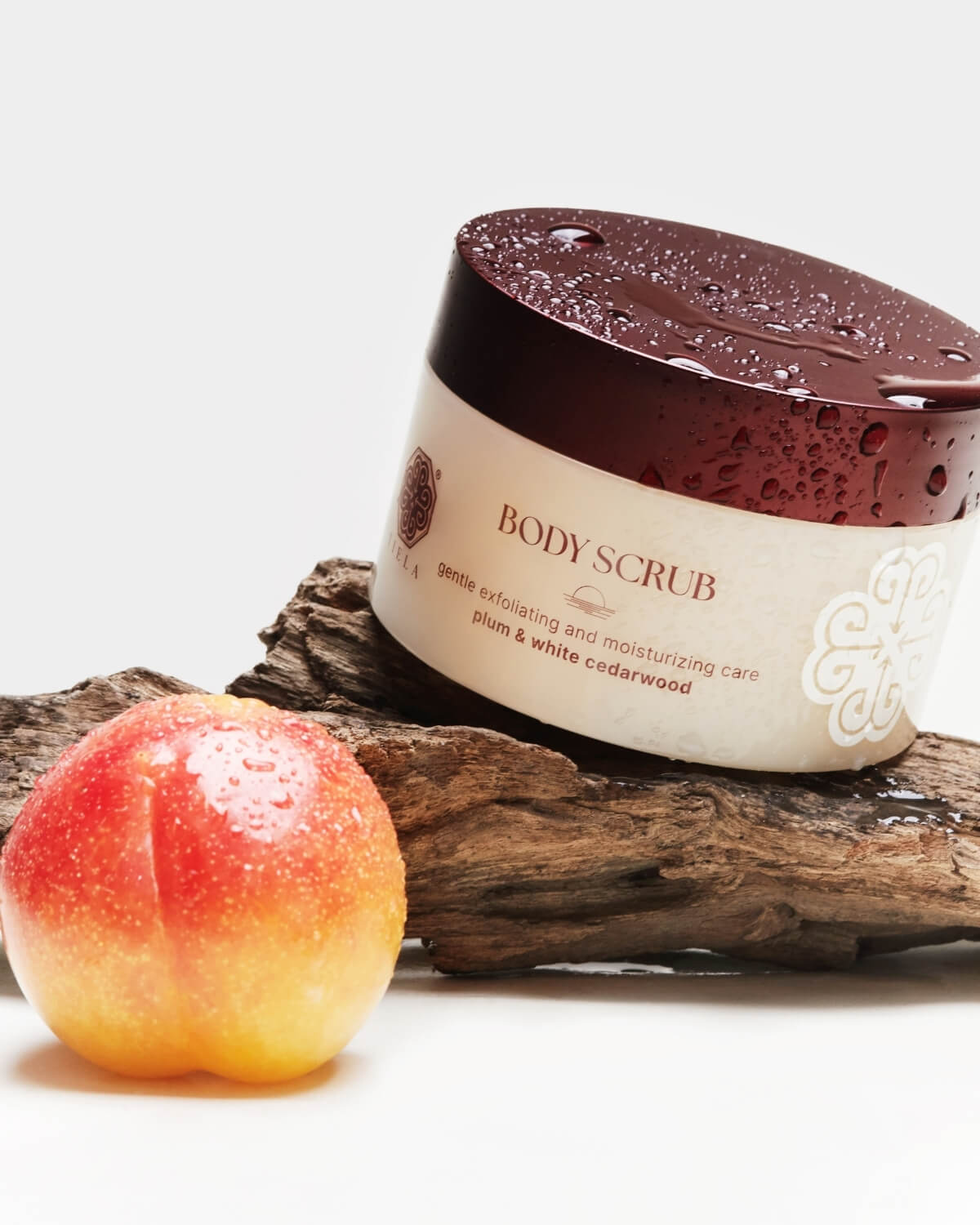 Tiela Body Scrub jar with a deep red, wet lid, balanced on a piece of dark driftwood next to a whole, wet plum.