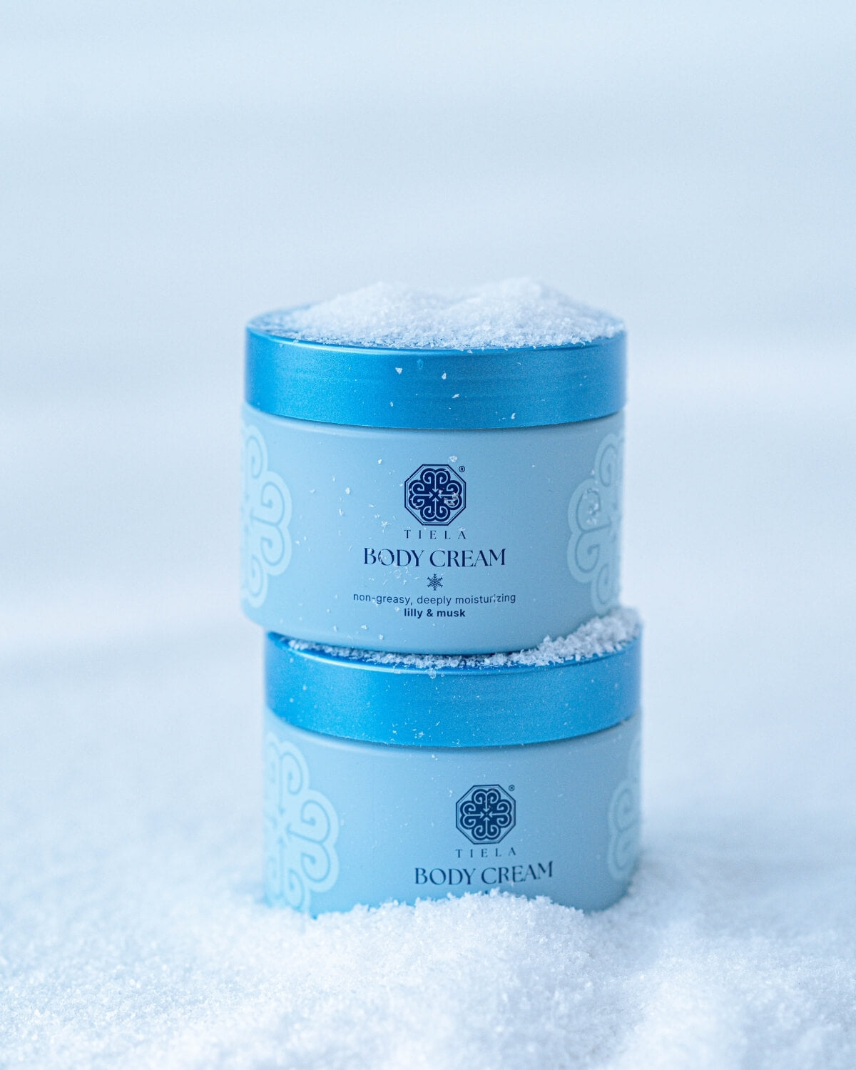 Two light blue Tiela Body Cream jars stacked and partially buried in white, crystalline material resembling snow or salt.
